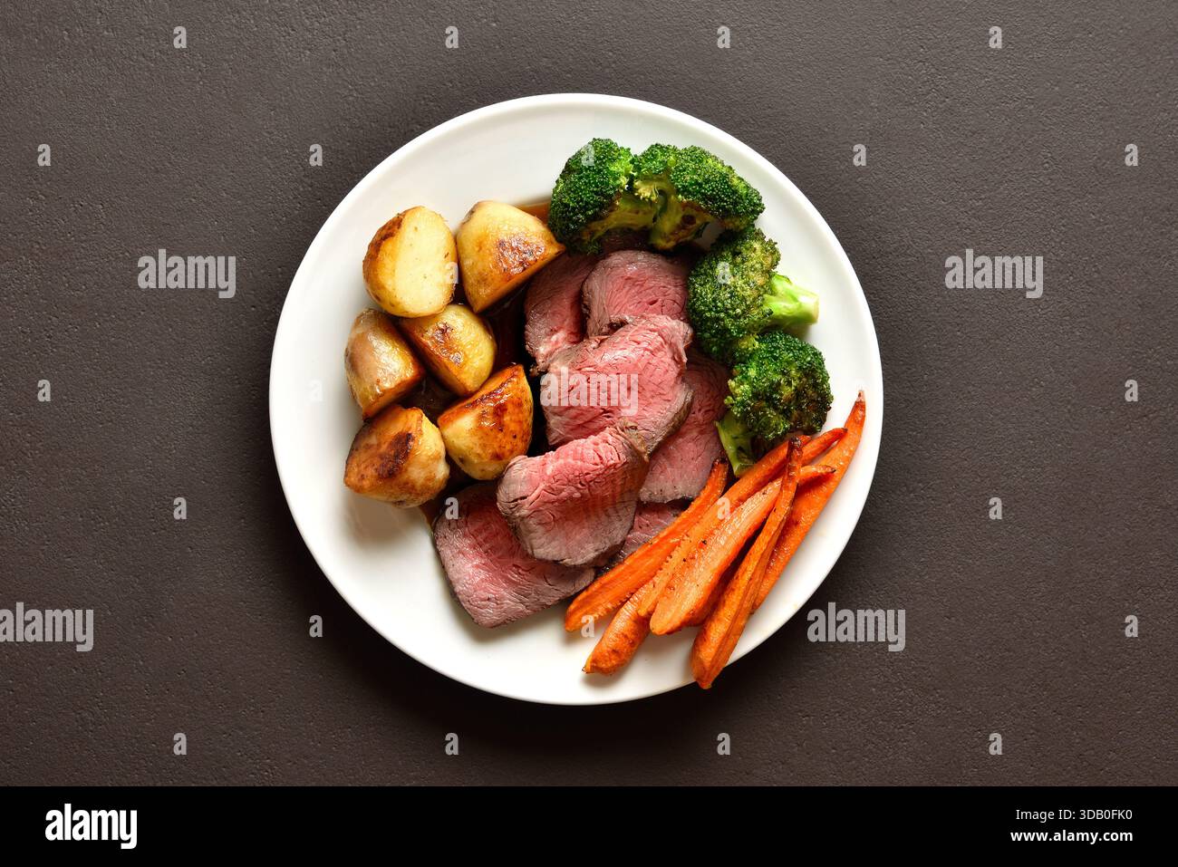 Sonntagsbraten mit gebratenem Rindfleisch, Karotten, knuspriger Kartoffel und Brokkoli auf weißer Platte auf braunem Steinhintergrund. Traditionelles britisches Gericht. Draufsicht, flach Stockfoto