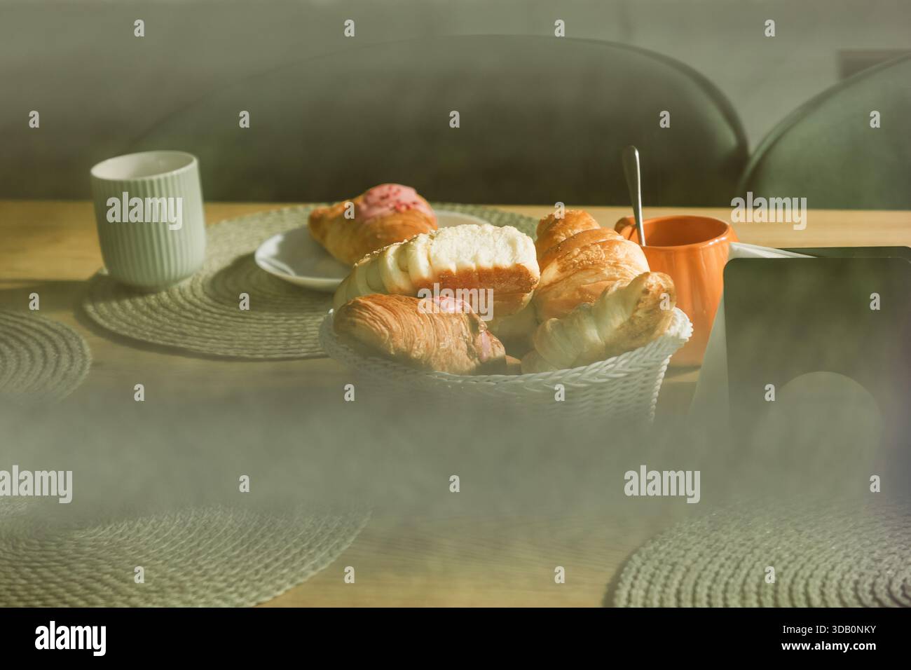 Eine warme, einladende Frühstücksszene mit einem Korb mit Croissants und Gebäck, einem weißen Becher und einer orangen Tasse auf einem Holztisch, was auf ho hindeutet Stockfoto