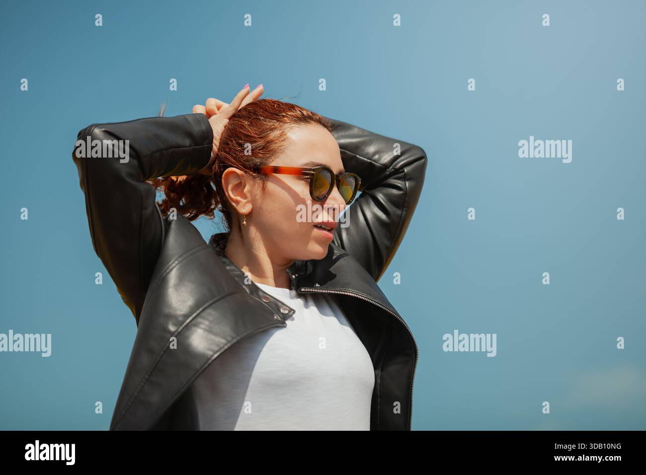 Rothaarige Frau in Sonnenbrille und Lederjacke, weißes Hemd, zieht die Haare zurück gegen den blauen Himmel und schaut zur Seite. Stockfoto