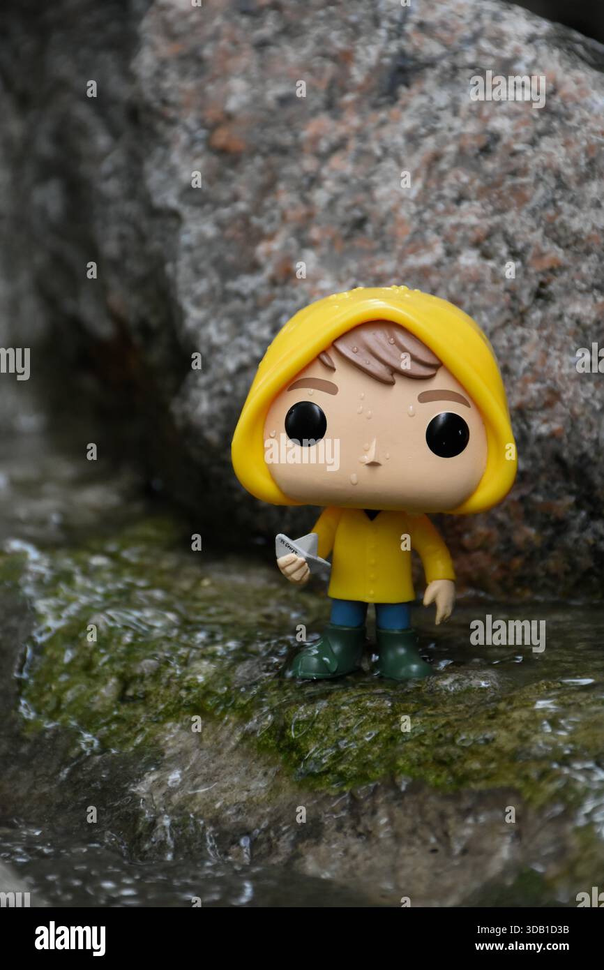 Funko Pop Actionfigur von Georgie Denbrough aus dem Horrorfilm IT. Junge mit Papierboot steht zwischen Felsen am Fluss. Stockfoto