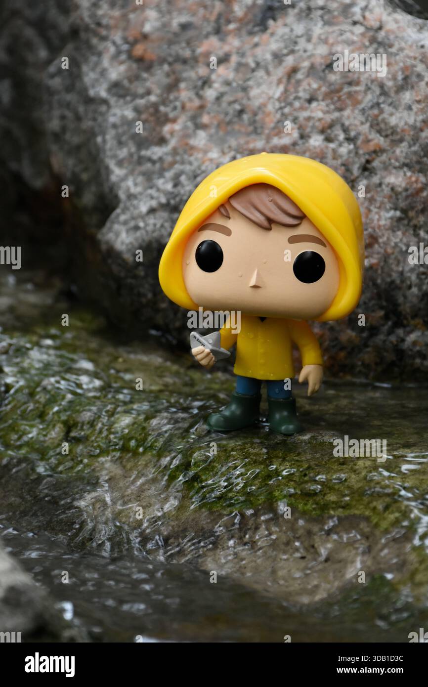 Funko Pop Actionfigur von Georgie Denbrough aus dem Horrorfilm IT. Junge mit Papierboot steht zwischen Felsen am Fluss. Stockfoto