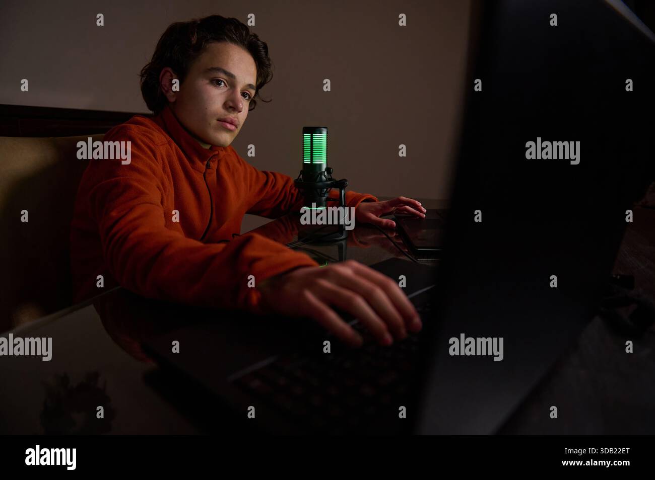 Ein junger Mann in einem orangefarbenen Hoodie sitzt an einem Schreibtisch mit einem Laptop und einem leuchtend grünen Mikrofon, fokussiert auf den Bildschirm. Gemütliches Studio für Streaming, Gami Stockfoto