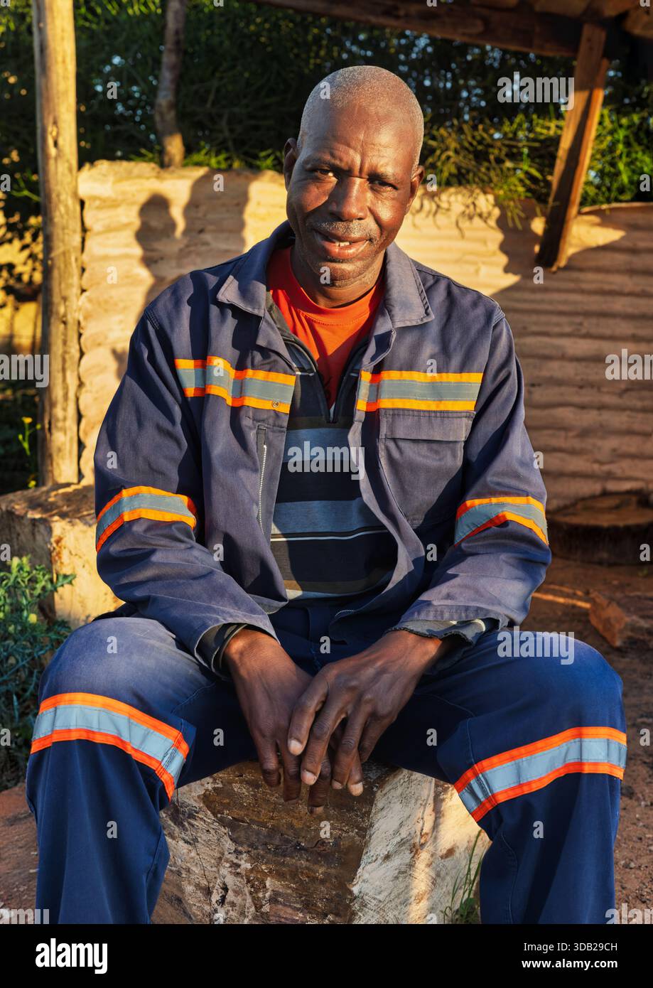 Einzelne afrikanische Männer sitzen draußen auf einer Steinbank bei Sonnenaufgang, Einstellung mit gewellten Metall- und Holzstützen, Johannesburg, Südafrika, blauer wo Stockfoto