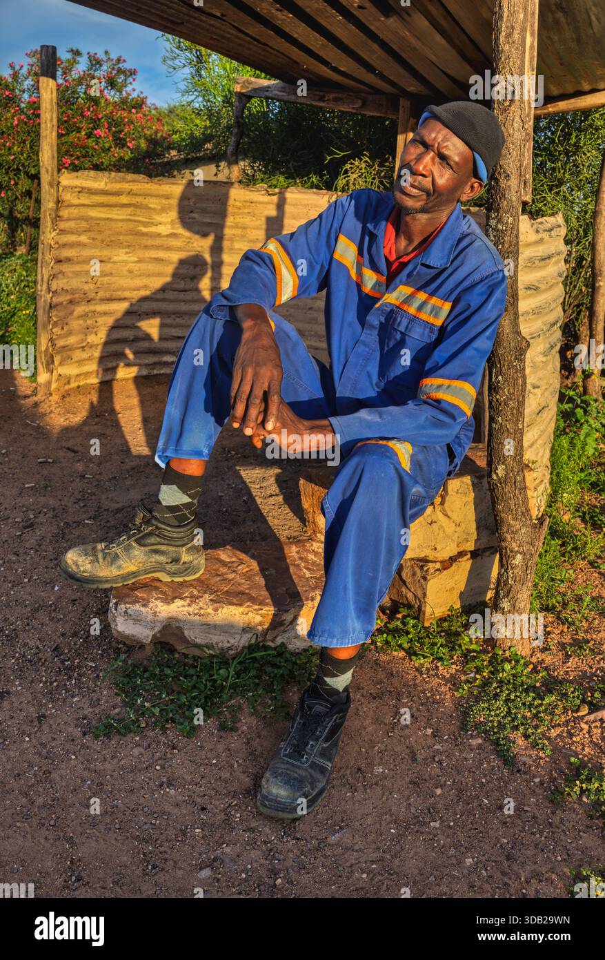 Einzelner afrikanischer Arbeiter sitzt draußen auf einer Steinbank bei Sonnenaufgang, Untergang hat gewellte Metall- und Holzstützen, Johannesburg, Südafrika, Stockfoto