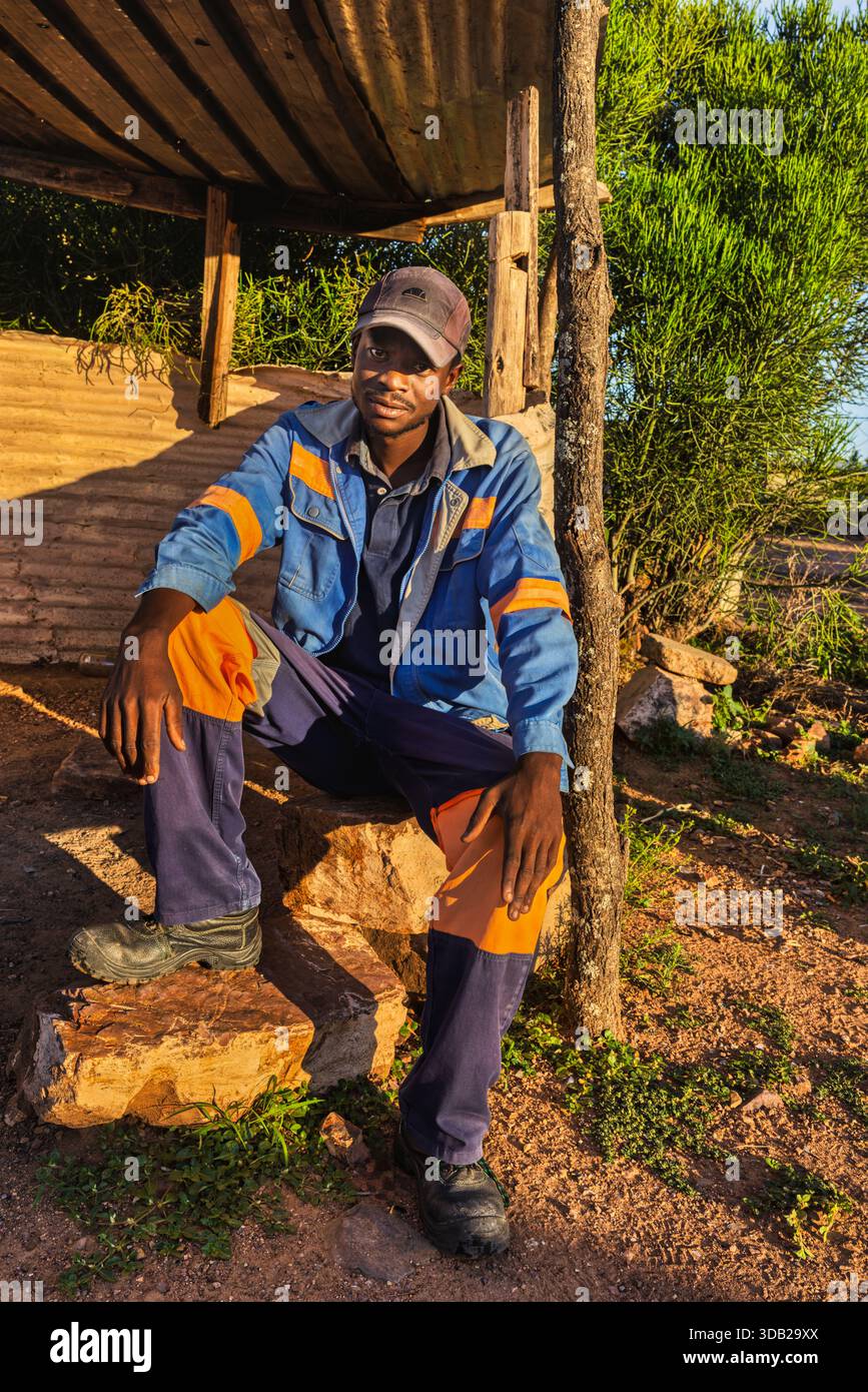 Einzelner afrikanischer Arbeiter sitzt draußen auf einer Steinbank bei Sonnenaufgang, Untergang hat gewellte Metall- und Holzstützen, Johannesburg, Südafrika, Stockfoto