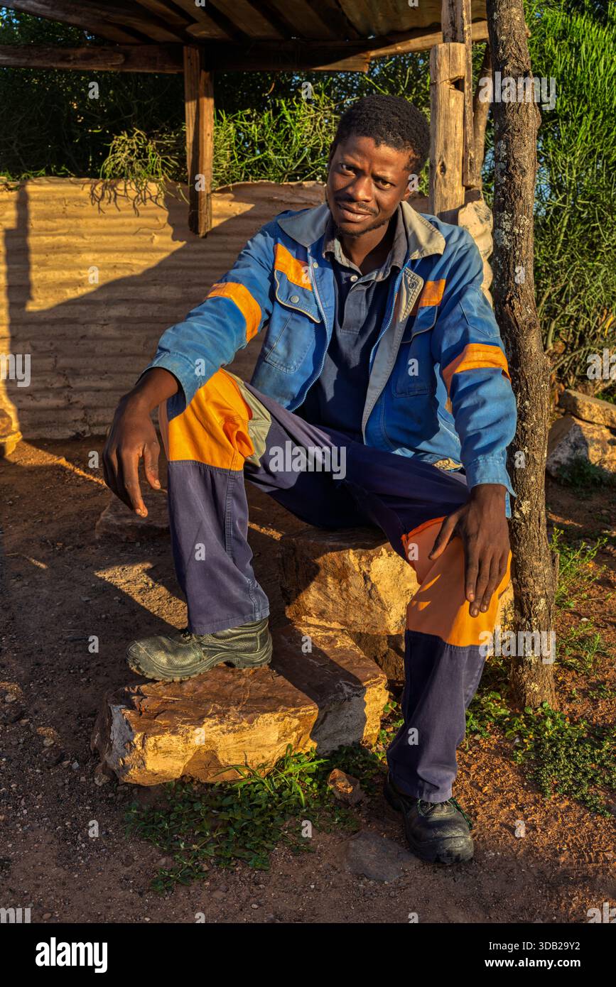 Einzelner afrikanischer Arbeiter sitzt draußen auf einer Steinbank bei Sonnenaufgang, Untergang hat gewellte Metall- und Holzstützen, Johannesburg, Südafrika, Stockfoto