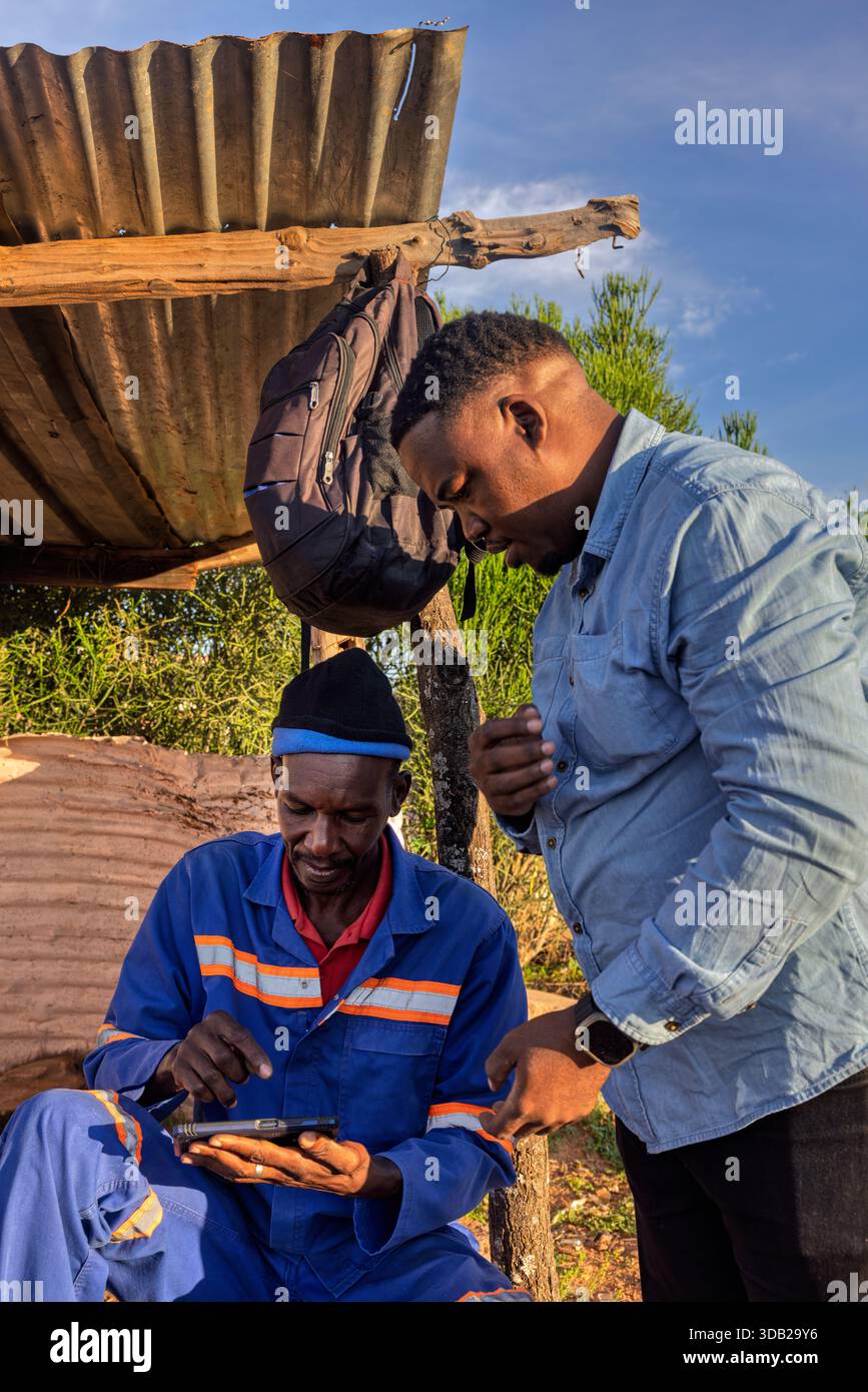 Zwei afrikanische Männer, die bei Sonnenaufgang draußen auf einer Steinbank sitzen, mit gewellten Metall- und Holzstützen, Johannesburg, Süd-Afri Stockfoto