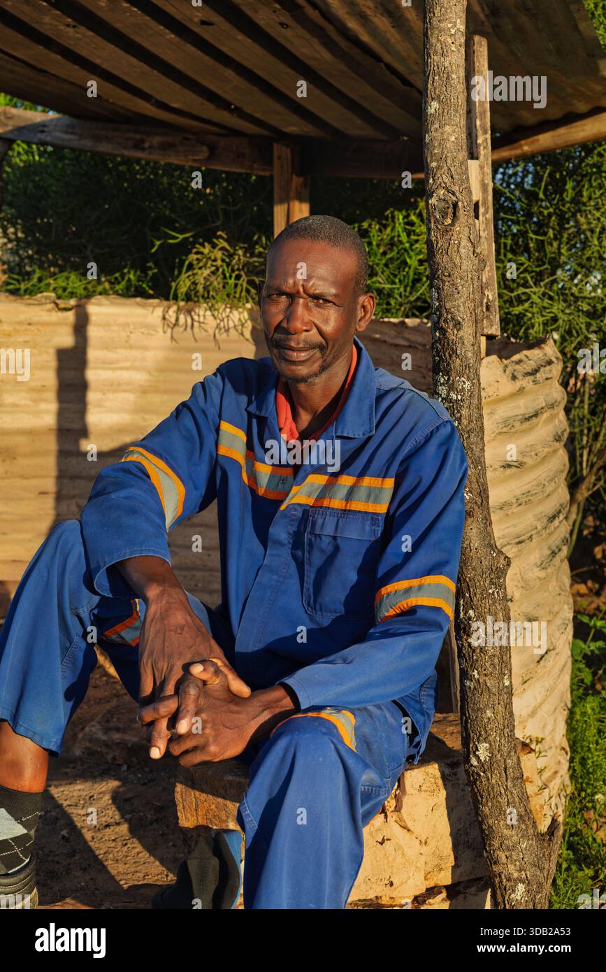 Einzelner afrikanischer Arbeiter sitzt draußen auf einer Steinbank bei Sonnenaufgang, Untergang hat gewellte Metall- und Holzstützen, Johannesburg, Südafrika, Stockfoto