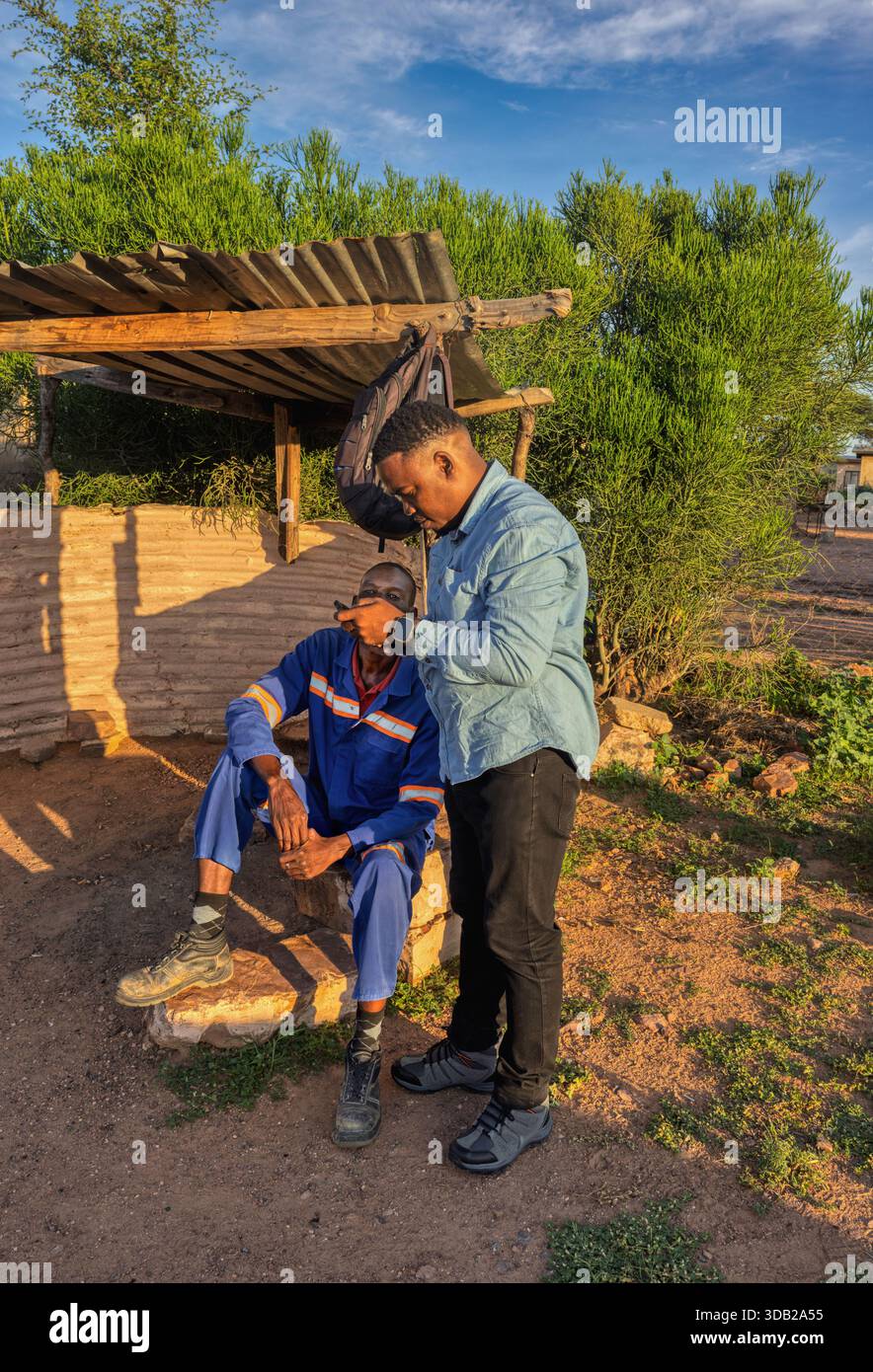 Zwei afrikanische Männer, die ein Telefon benutzen, sitzen draußen auf einer Steinbank bei Sonnenaufgang, der Hintergrund hat gewellte Metall- und Holzstützen, Johannesburg, South AFR Stockfoto