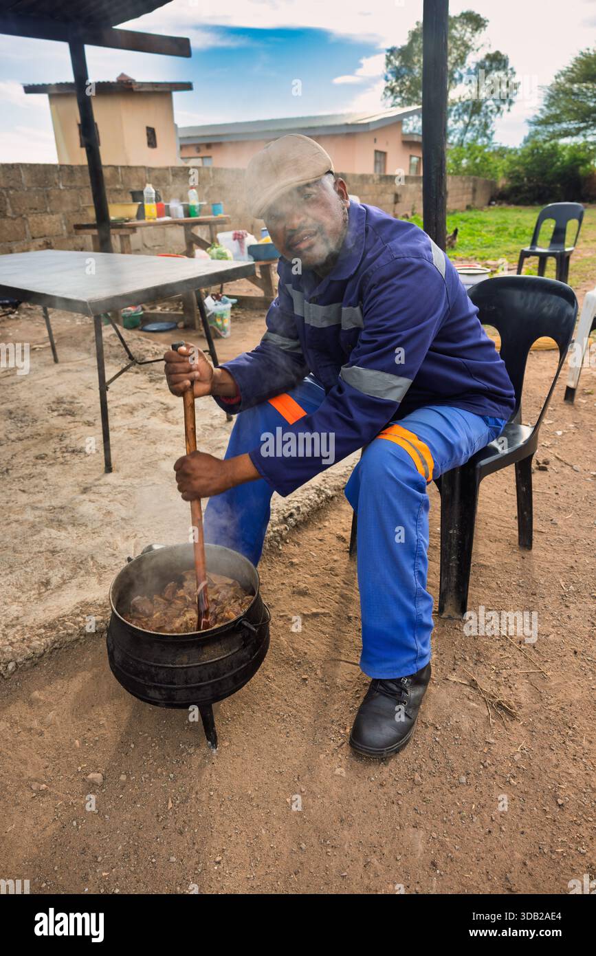 afrikanische Männer, die im Freien Seswaa in einem dreibeinigen gusseisernen Topf zubereiten, für Eintöpfe oder Potjiekos, in Arbeitskleidung, Botswana, Südliches Afrika Stockfoto