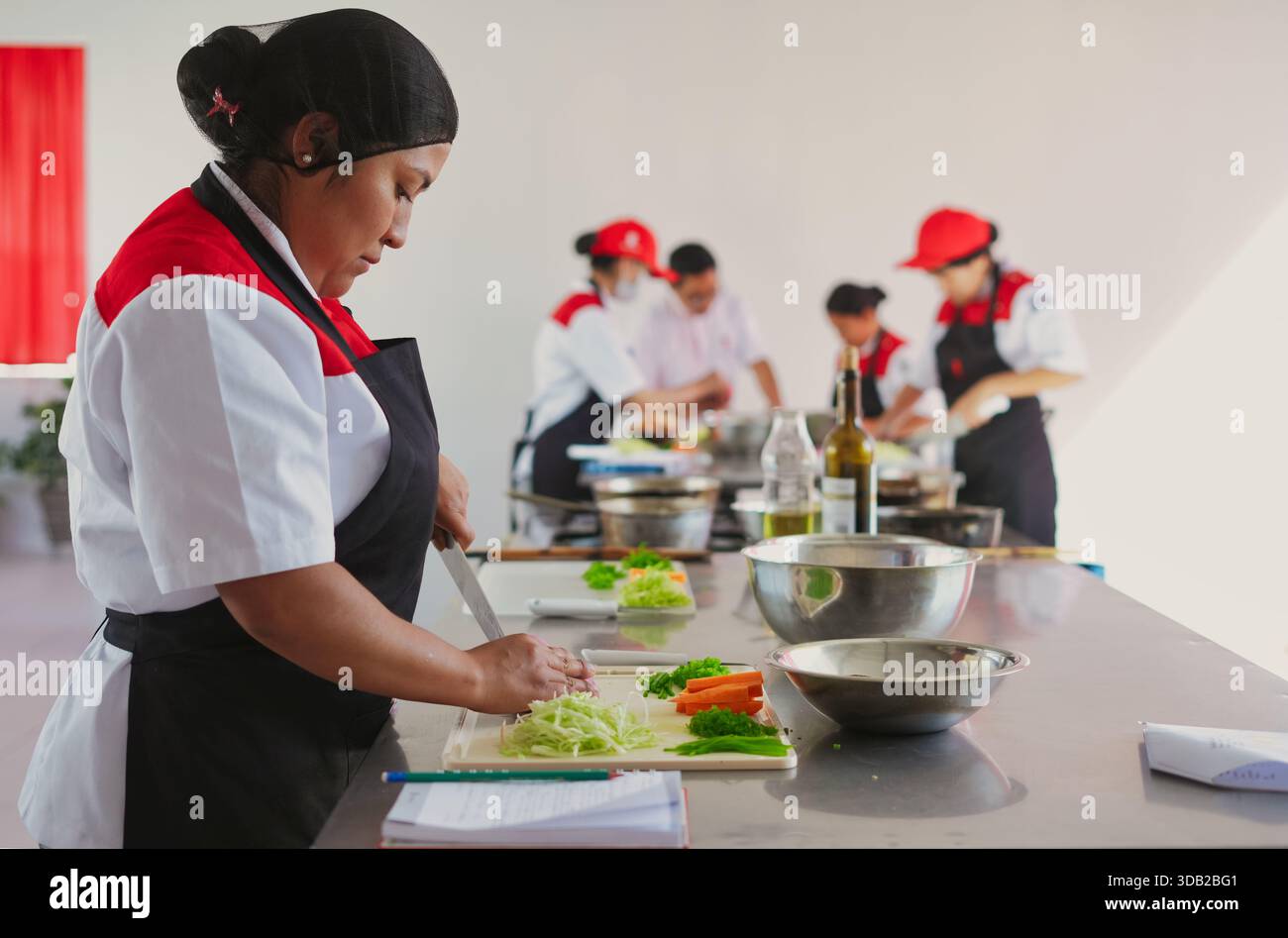 Küchenchefin, die frisches Gemüse auf einem Schneidebrett hackt, zusammen mit Mitschülern in einer professionellen Küche kulinarische Fähigkeiten erlernt, Focus Stockfoto