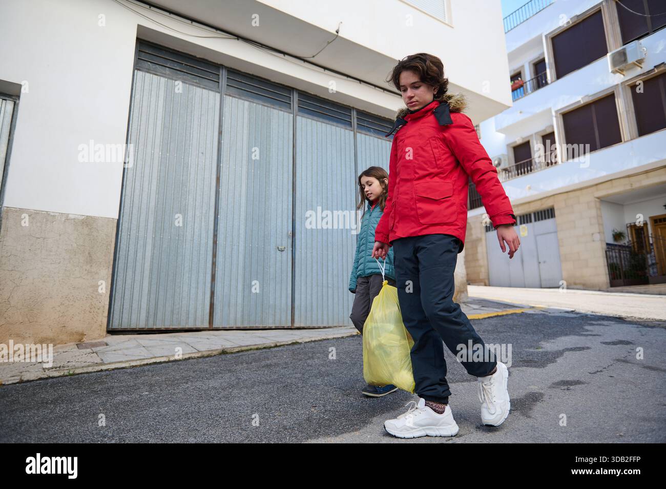Zwei Kinder in Jacken laufen auf einer sonnigen Straße entlang und tragen eine gelbe Plastiktüte. Eine einfache Szene des Alltags und Familienausflugs in einer Wohnung Stockfoto