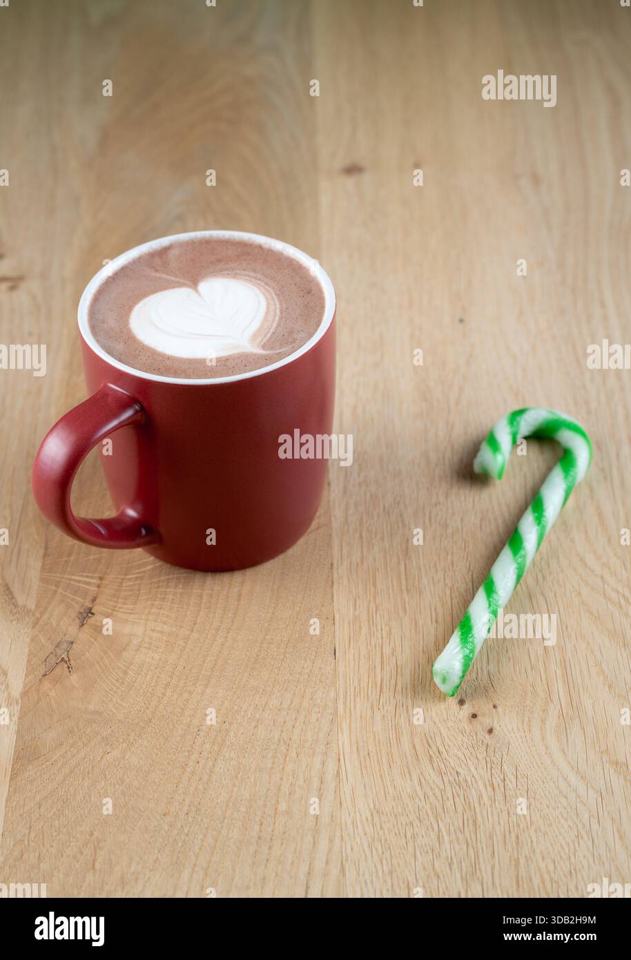 Ein roter Becher mit heißer Schokolade mit einem Herzen darauf sitzt auf einem Holztisch. Ein grün-weiß gestreifter Zuckerrohr ist neben dem Becher Stockfoto
