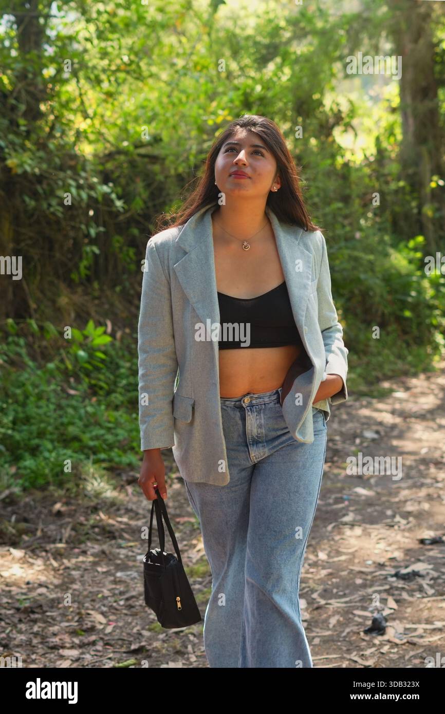 Junge Erwachsene Frau, die einen sonnendurchfluteten Feldweg in einem grünen Wald entlang geht und mit einem besinnlichen Ausdruck aufblickt, während sie eine Handtasche trägt und trägt Stockfoto