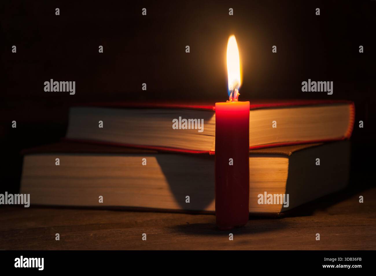 Eine rote Kerze brennt auf einem Holztisch, ihr warmes Leuchten beleuchtet zwei Bücher dahinter. Eine sanfte, ruhige Atmosphäre, ideal für Themen wie Lesen, Gemütlichkeit Stockfoto