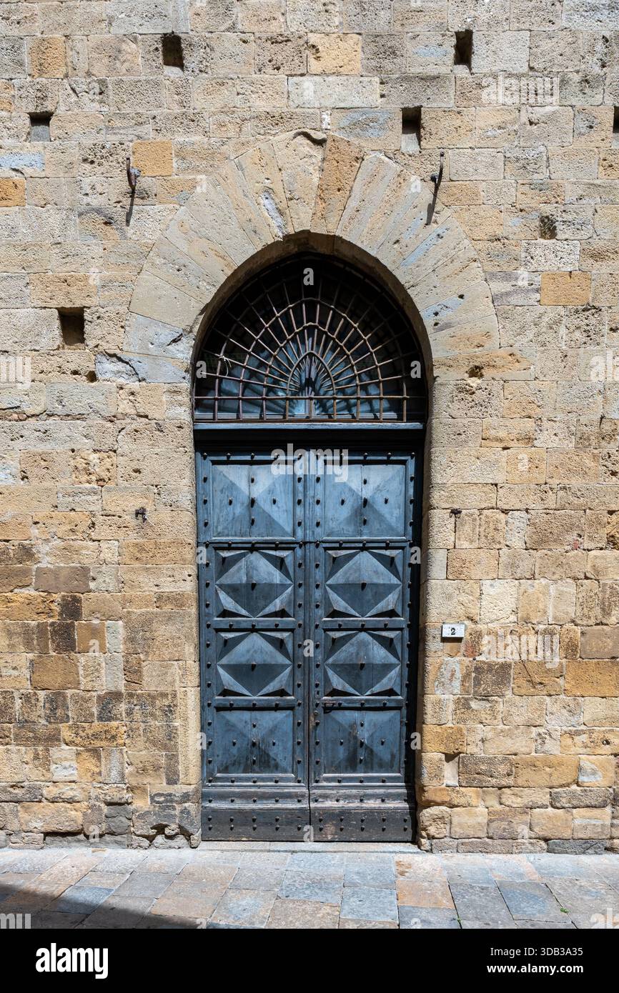 Mittelalterliches Steinportal des Palazzo dei priori in Volterra, stabile Eisentür unter spitzem Bogen auf der sonnendurchfluteten piazza. Stockfoto