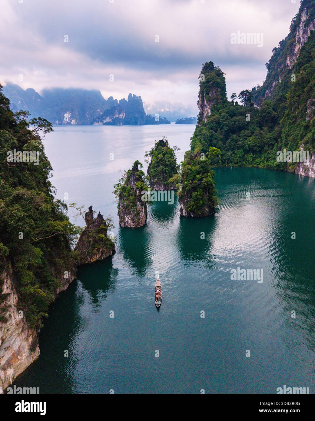 Erkunden Sie das ruhige Wasser des Cheow Lan Lake, umgeben von üppigem Grün und hoch aufragenden Kalksteinklippen. Ein einsames Boot gleitet durch die ruhige Landschaft, Khao Sok Thailand Stockfoto