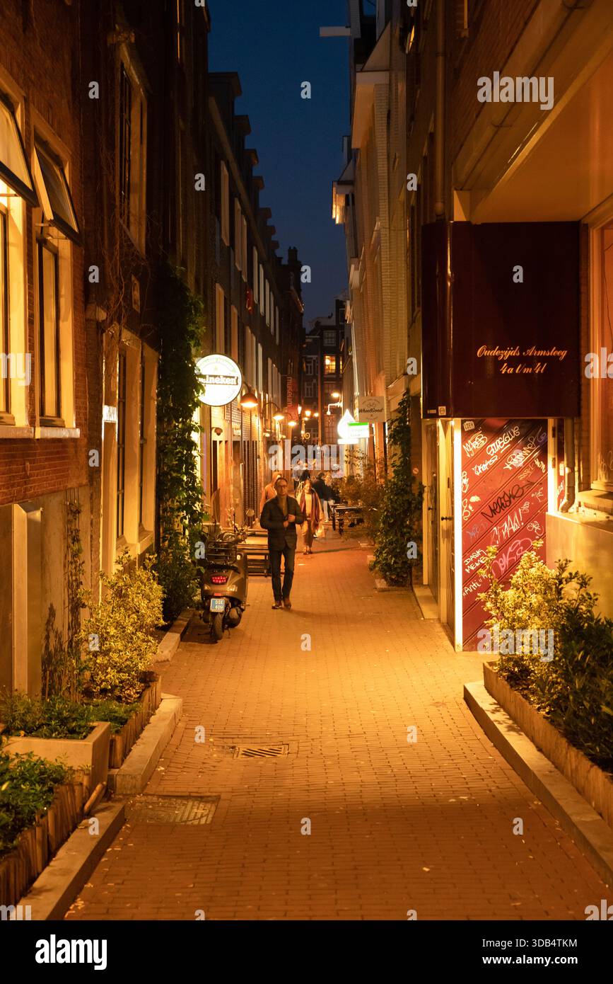 Warme, schmale Gasse in Amsterdam bei Nacht mit Fußgängern, geparkten Rollern und Graffiti Stockfoto