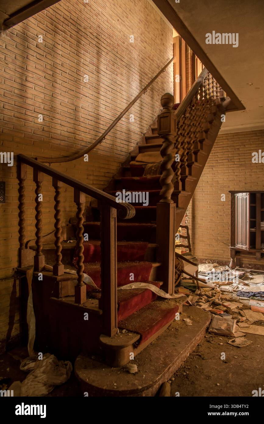 Verstaubte geschwungene Treppe mit rotem Teppich in einer verlassenen Villa mit verzierten, verdrehten Balustern und verstreuten Trümmern Stockfoto