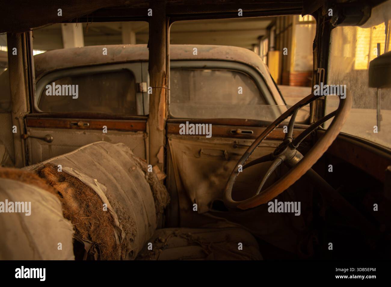 Verrostetes Oldtimer-Interieur mit abgerissener Polsterung und Holzlenkrad in einer verlassenen Garage Stockfoto