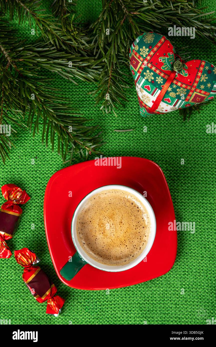 Eine Tasse Kaffee mit schaumiger Crema in einer dunkelgrünen Tasse auf einer roten Untertasse. Der Hintergrund ist mit immergrünen Zweigen und einem herzförmigen Ornament versehen Stockfoto