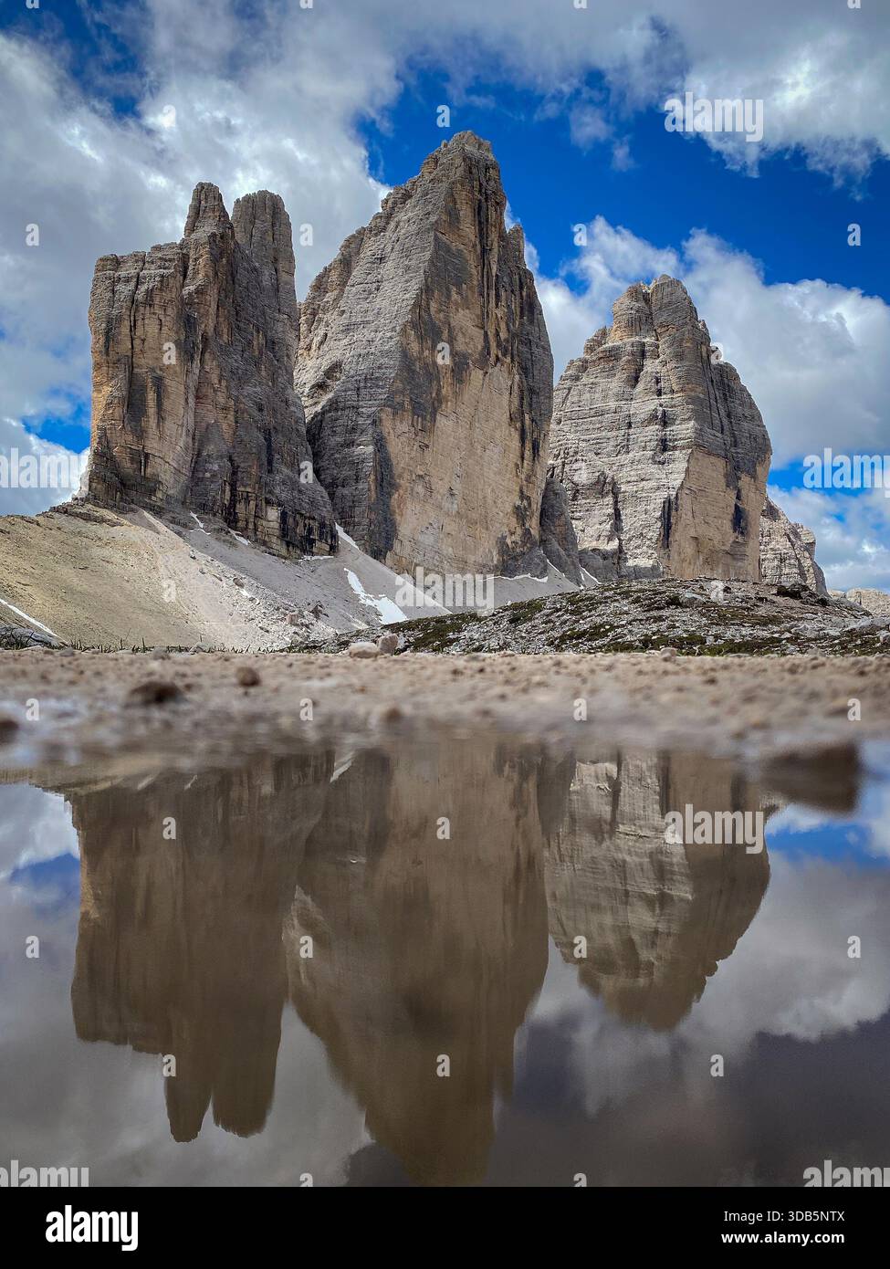 Niedriger Blickwinkel auf Tre Cime di Lavaredo in einer Pfütze; Cima Grande, Cima Ovest und Cima Piccola über hellem Kalksteingeröll vor blauem Himmel mit Wolken Stockfoto