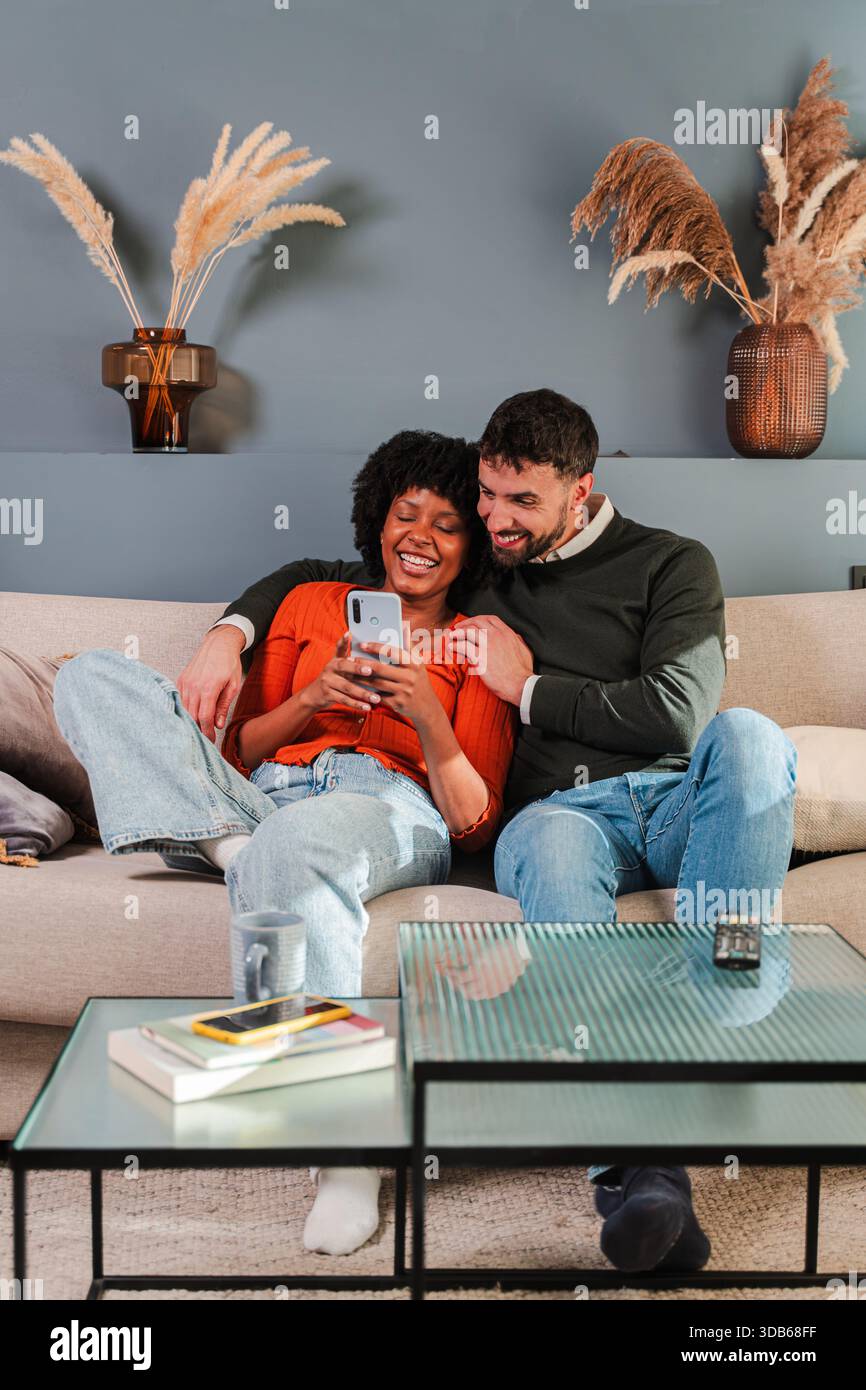 Ein entzücktes Paar genießt eine gute gemeinsame Zeit, während sie lachen, während sie ein Smartphone auf der Couch verwenden. Sie sind motiviert und inhaltlich, kreativ Stockfoto