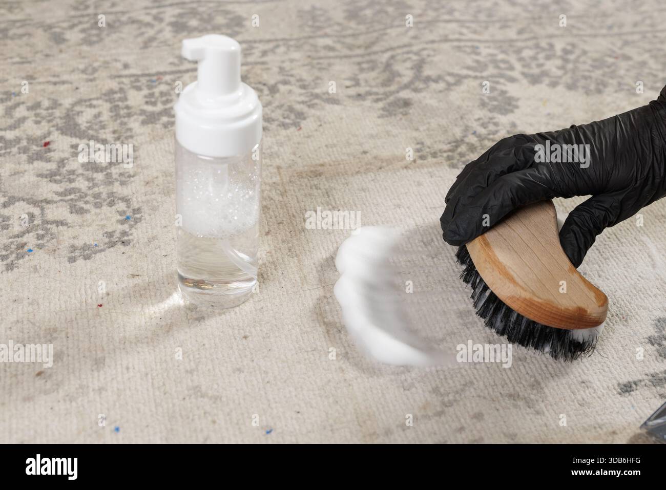 Arbeiter in schwarzen Handschuhen verteilt Schaumreiniger auf Vintage-Teppichoberfläche mit Scheuerbürste und Flasche in der Nähe Stockfoto