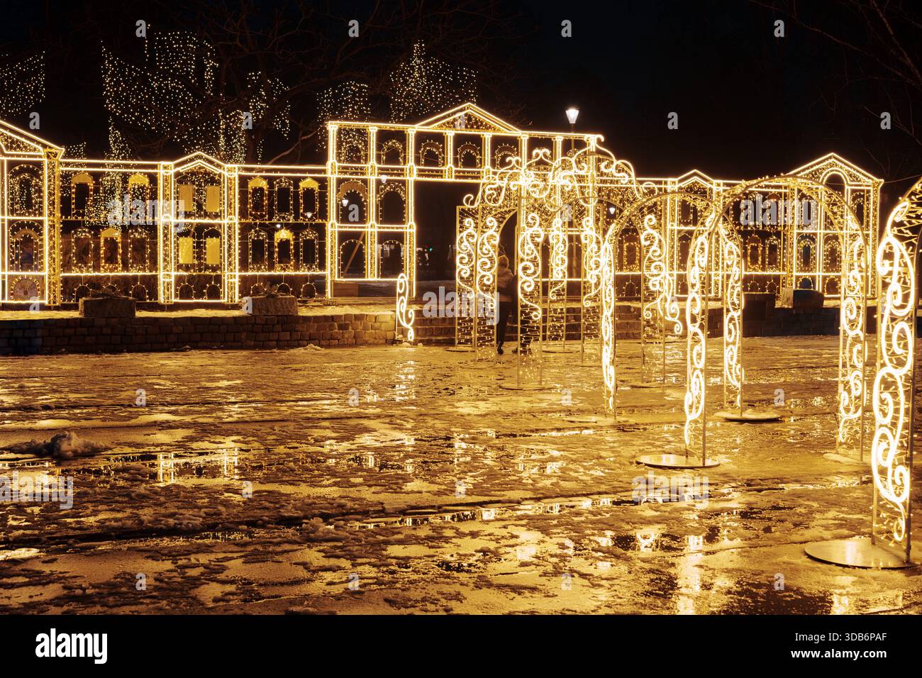 Festlicher Gehweg-Tunnel aus goldenen Weihnachtslichtbögen. Urbane Nachtkulisse mit leuchtenden Dekorationen, die sich auf nassem Boden spiegeln. Stockfoto