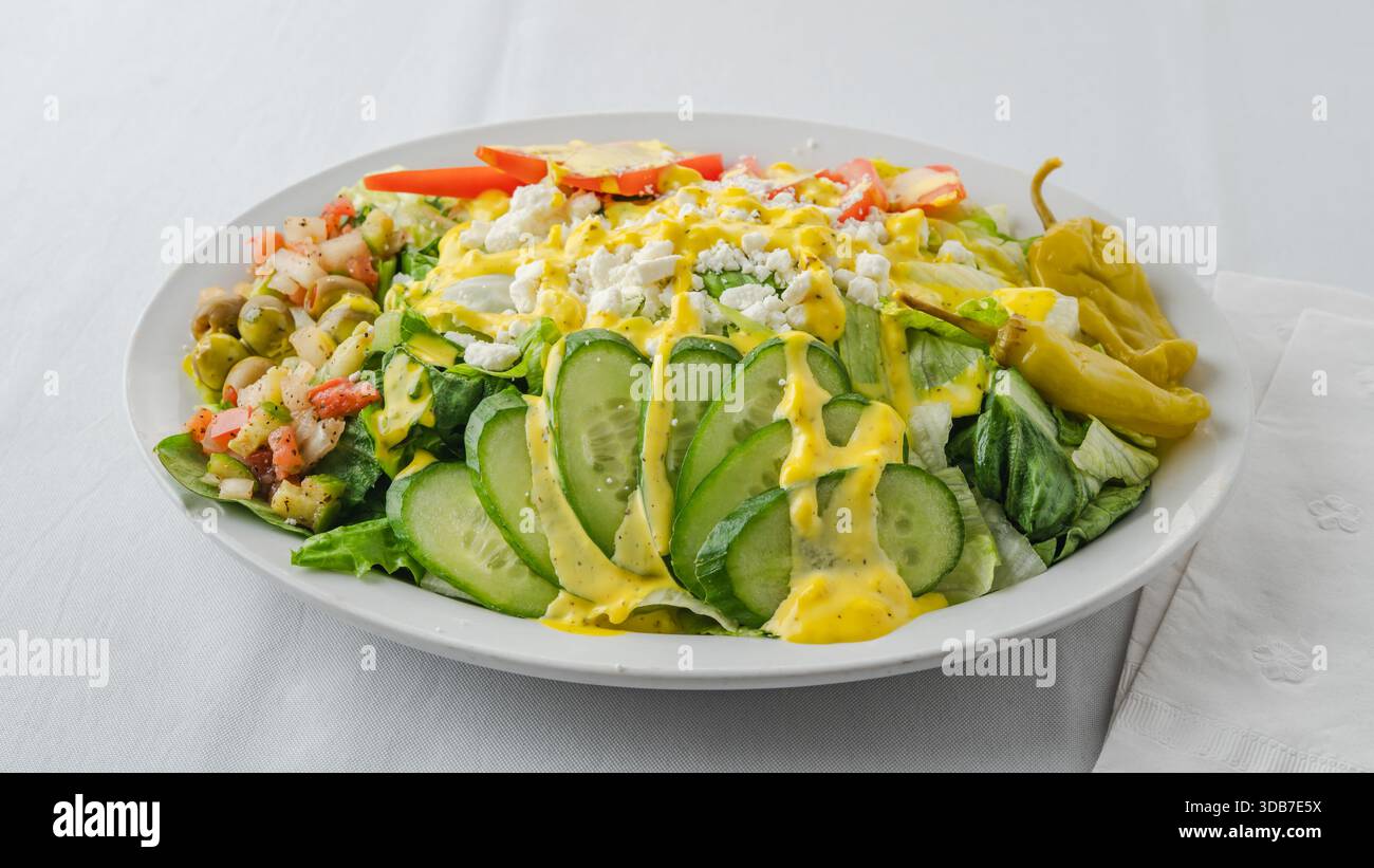 Ein lebendiger Salat mit Gurkenscheiben, Tomaten, Fetakäse und gelbem Dressing. Stockfoto
