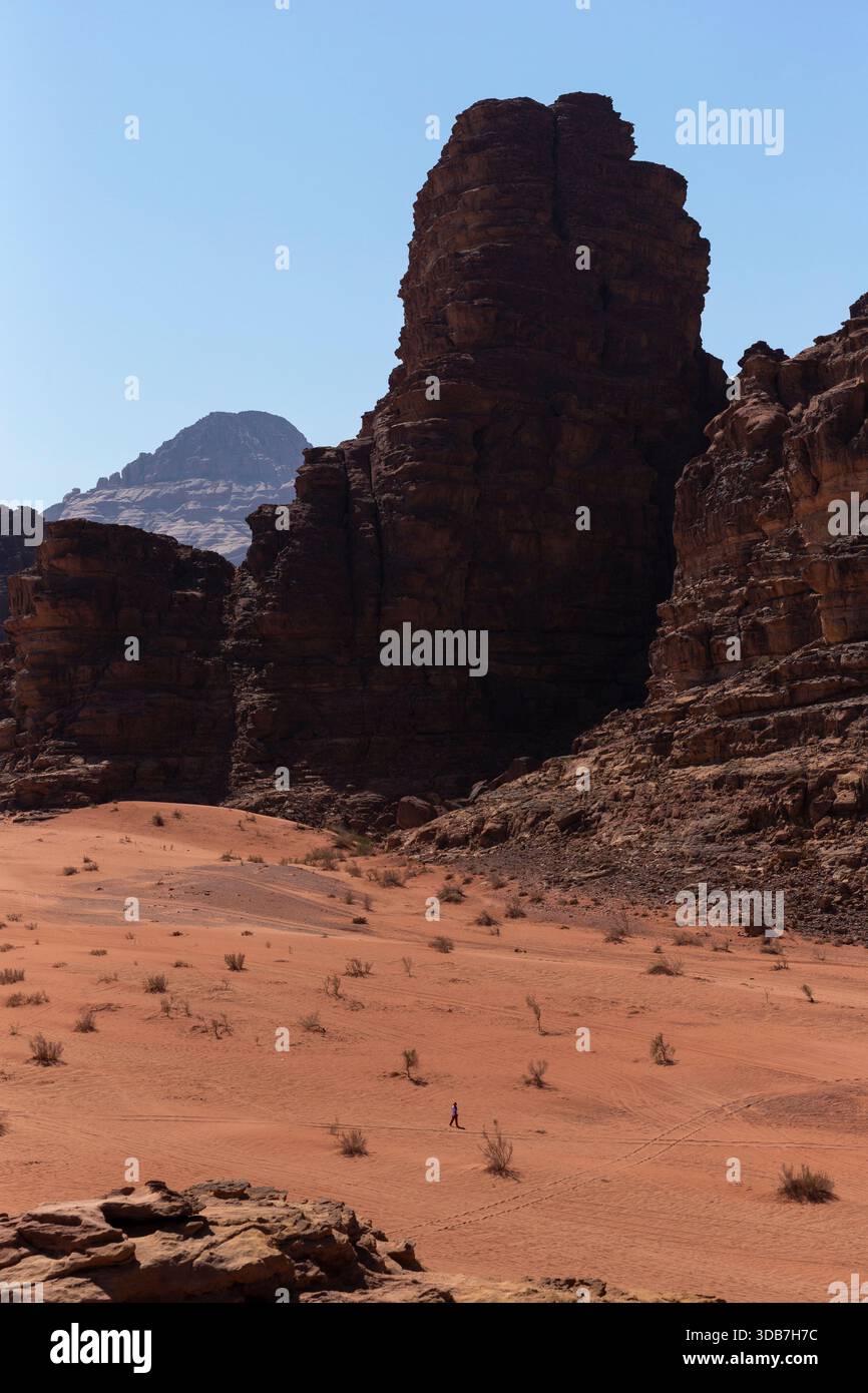 Eine hohe Sandsteinformation erhebt sich über einem weiten Wüstenboden in Wadi Rum, Jordanien, mit einer kleinen Person, die über den Sand läuft. Stockfoto