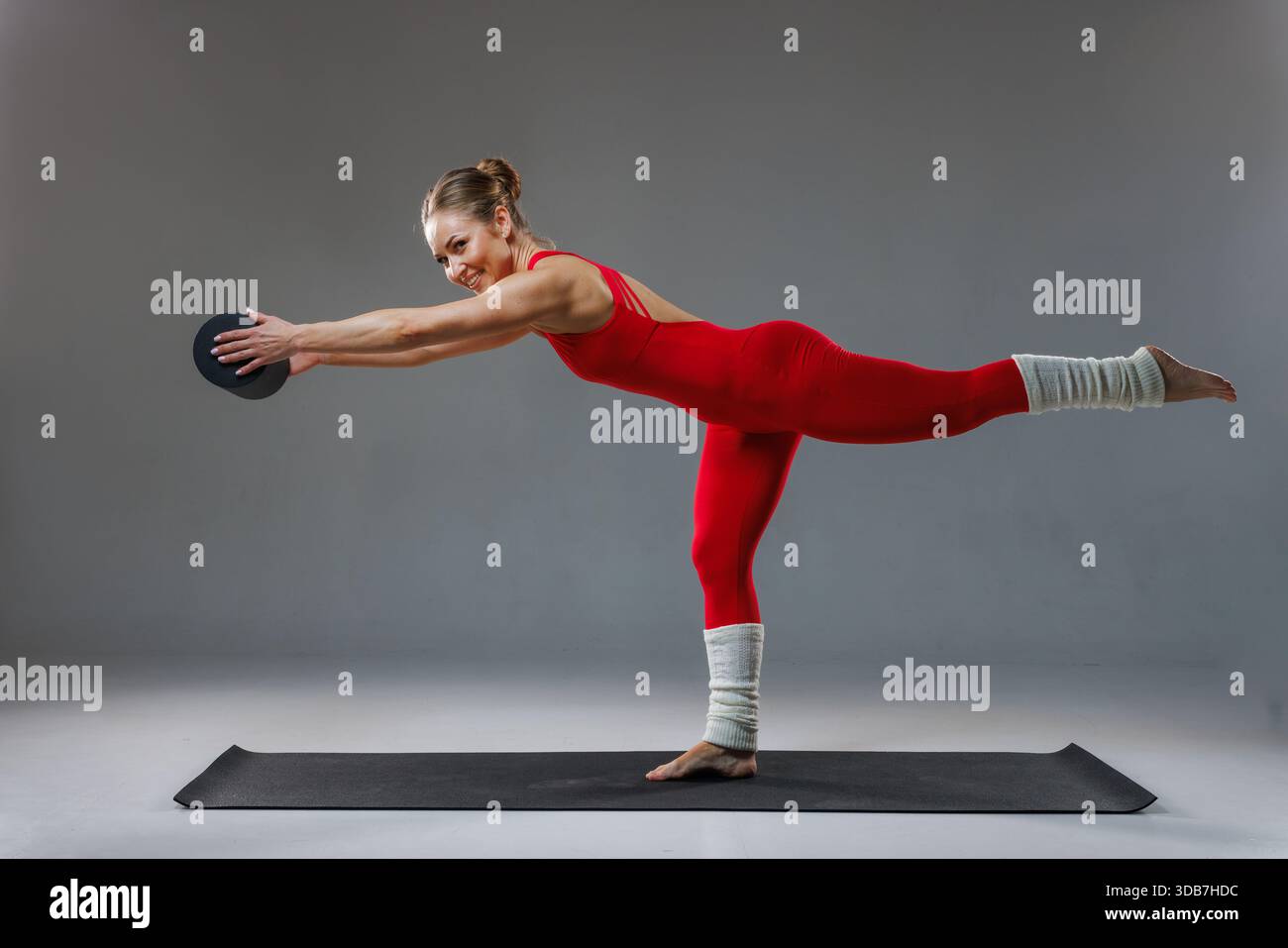 Fitte Frau, die Yoga in Warrior III-Pose übt, mit einem kleinen Gewicht, balanciert auf einem Bein, trägt rote Sportbekleidung und Beinstulpen auf einer Matte Stockfoto