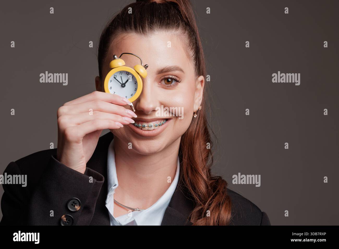 Lächelnde Geschäftsfrau mit Zahnspange, die einen kleinen gelben Wecker über dem Auge hält, Konzept des Zeitmanagements, Termine und Effizienz. Stockfoto