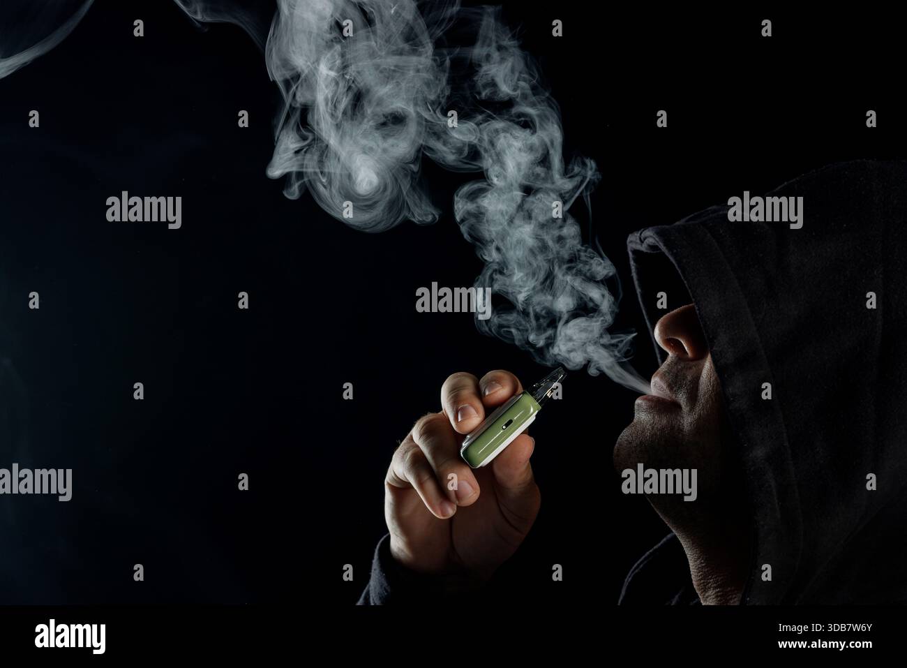 Geheimnisvoller Mann mit Kapuze, der mit E-Zigarette dampft, dichte Rauchwolken auf schwarzem Hintergrund, moderner Lebensstil und Gesundheitsrisiko bläst. Stockfoto