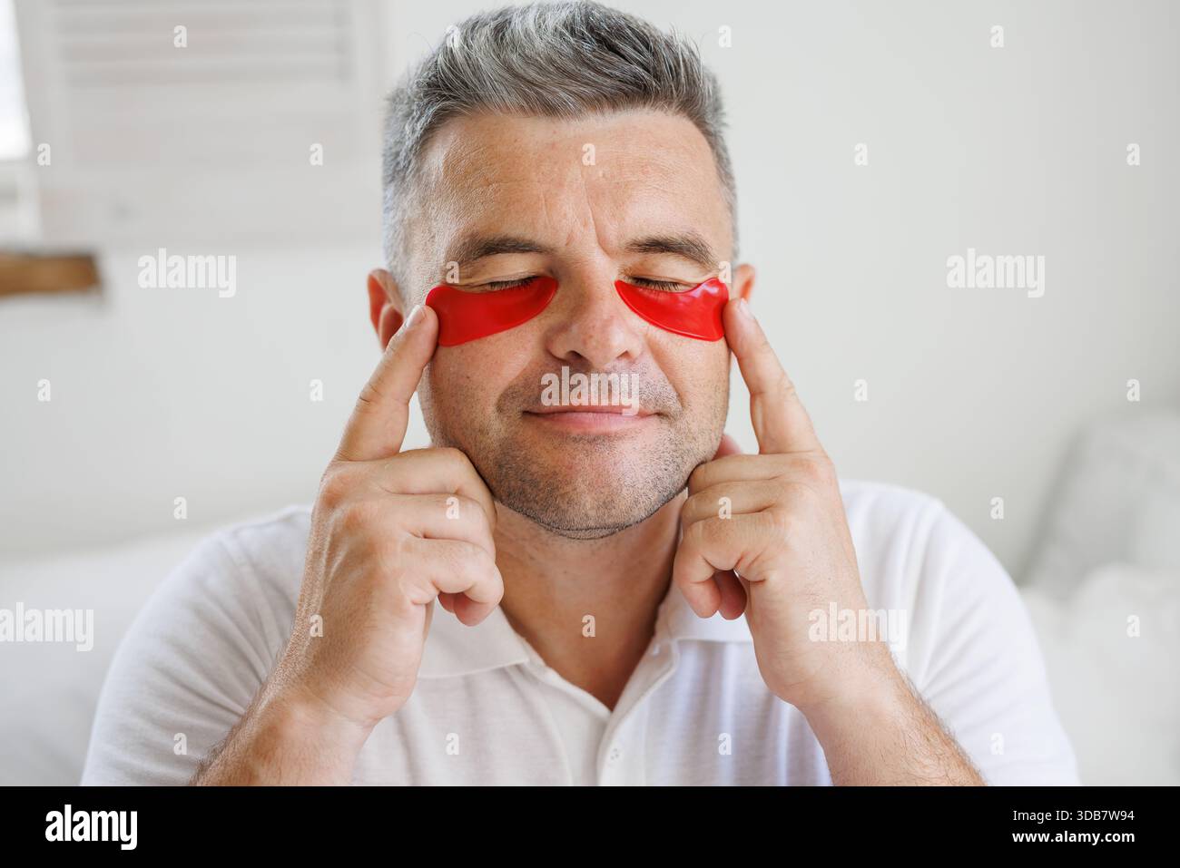 Ein lächelnder Mann mittleren Alters mit rotem Hydrogel unter den Augenflecken genießt Hautpflege, Verjüngung und Selbstpflege-Schönheitsbehandlungen zu Hause. Stockfoto