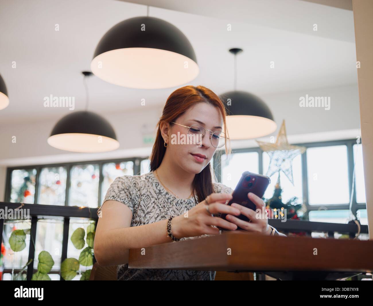 Die junge Frau benutzt ihr Smartphone, während sie an einem Tisch unter modernen Lichtern sitzt. Digitale Kommunikation und mobiles Lifestyle-Konzept. Stockfoto