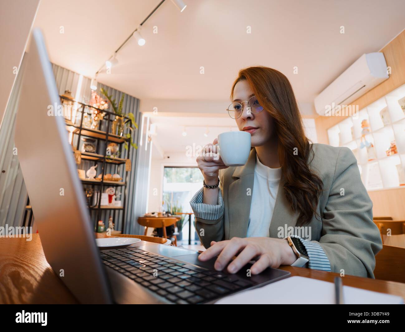 Professionelle Frau trinkt Kaffee, während sie einen Laptop an einem Kaffeetisch benutzt. Konzept von Pause, Produktivität und Remote-Arbeitsstil. Stockfoto