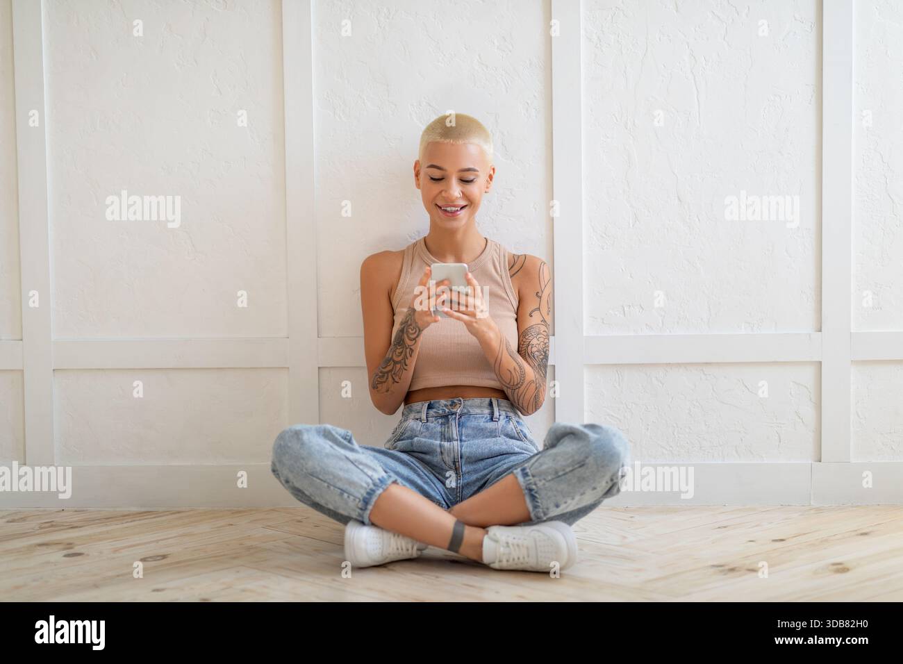 Glückliche Dame, die per Smartphone SMS schreibt und im Internet surft, auf dem Boden über der weißen Wand sitzt, Kopierraum Stockfoto