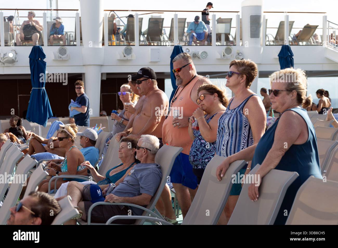 Kreuzfahrtpassagiere beobachten und reagieren auf Livemusik auf dem Pooldeck an Bord der Norwegian Encore und genießen während einer Tagesparty am Deck Getränke und Sonnenschein. Stockfoto