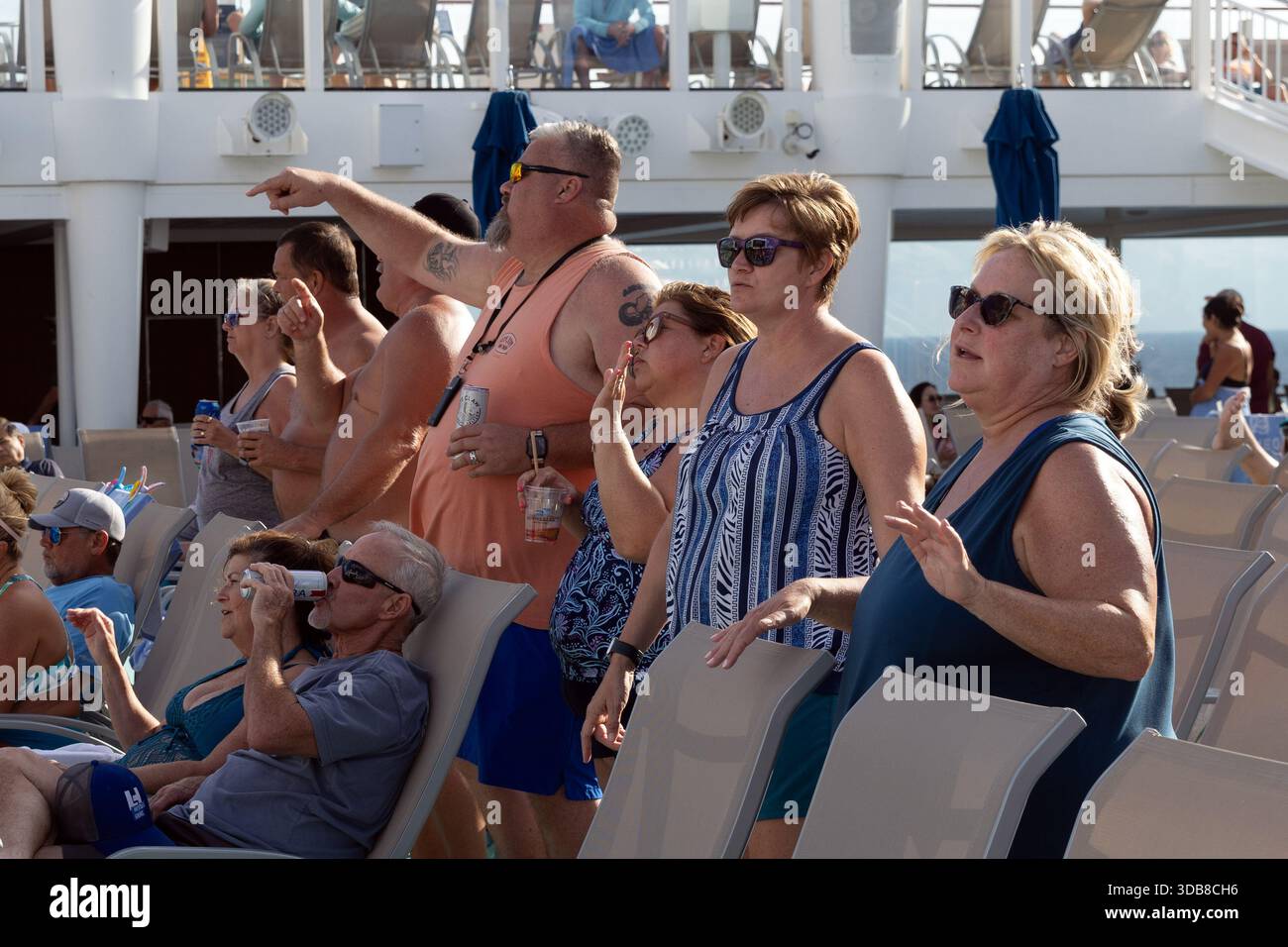Kreuzfahrtpassagiere beobachten und reagieren auf Livemusik auf dem Pooldeck an Bord der Norwegian Encore und genießen während einer Tagesparty am Deck Getränke und Sonnenschein. Stockfoto