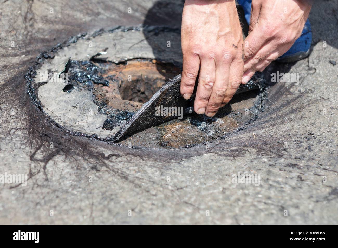 Der Arbeiter hebt bei der Reparatur des Flachdachdrainagesystems beschädigtes Dachmaterial auf und legt rostiges Metall und teerbeschichtete Schichten frei. Nahaufnahme markiert in Stockfoto