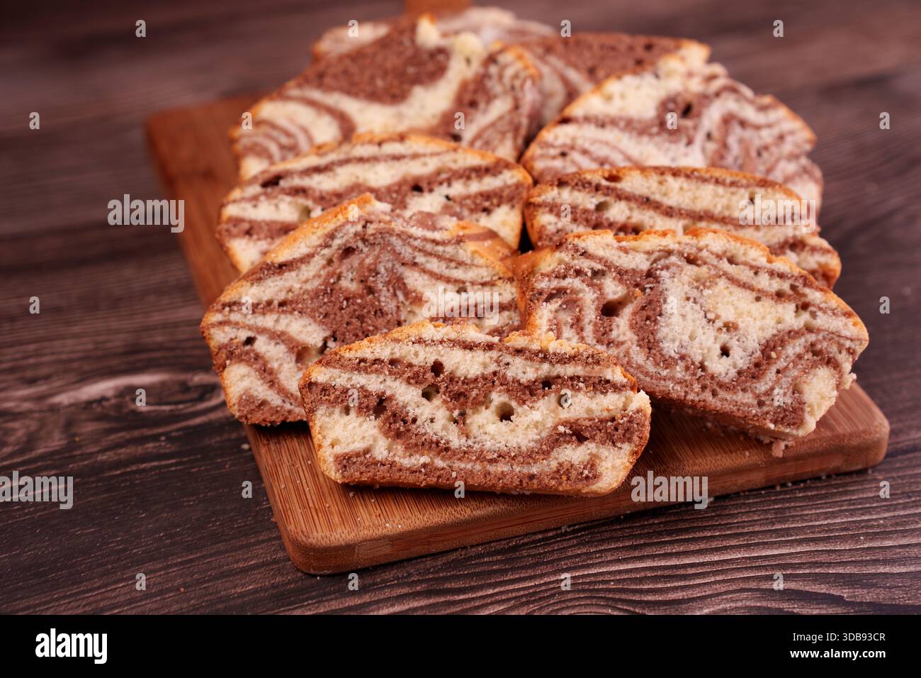 In Scheiben geschnittener Marmorkuchen mit Schokoladen- und Vanilleschmuck auf Holzbrett vor dunklem hölzernem Hintergrund, der Textur und Kontrast in einem gemütlichen Rost hervorhebt Stockfoto