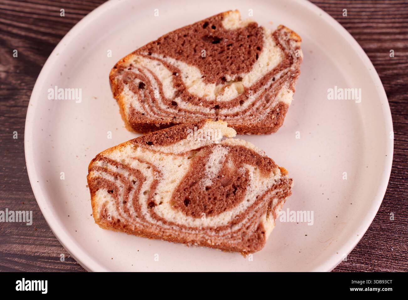 Nahaufnahme von zwei Scheiben Marmorkuchen mit einem wirbelnden Muster aus Schokolade und Vanille, die auf einem hellen Teller vor einem dunklen Holzboden ruhen Stockfoto