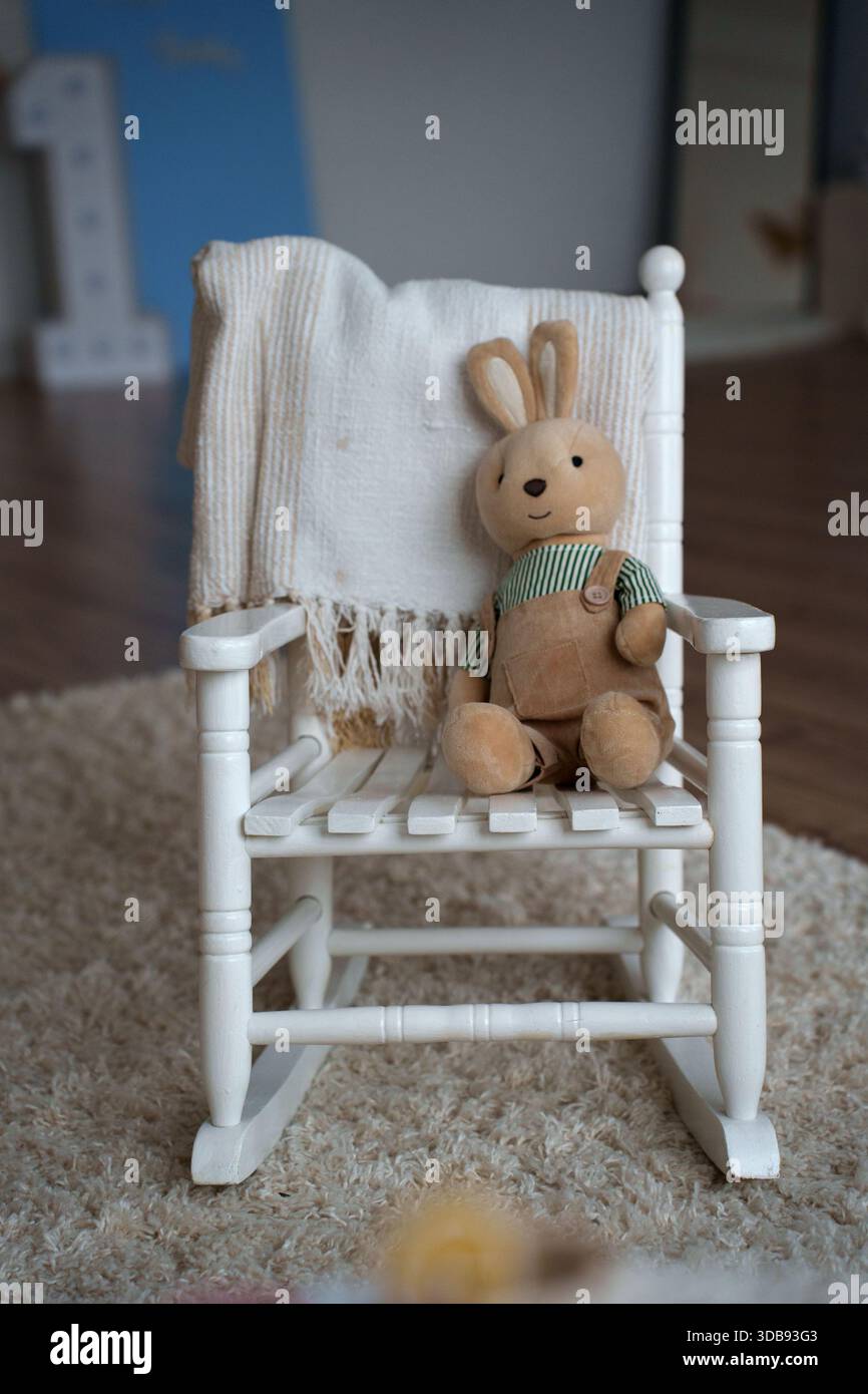 Gemütliche Szene mit entzückendem Plüschhasen im Schaukelstuhl aus Holz, mit weicher Decke auf flauschigem Teppich im ruhigen Kinderzimmer, Spiel, Kindheit, Unschuld Stockfoto