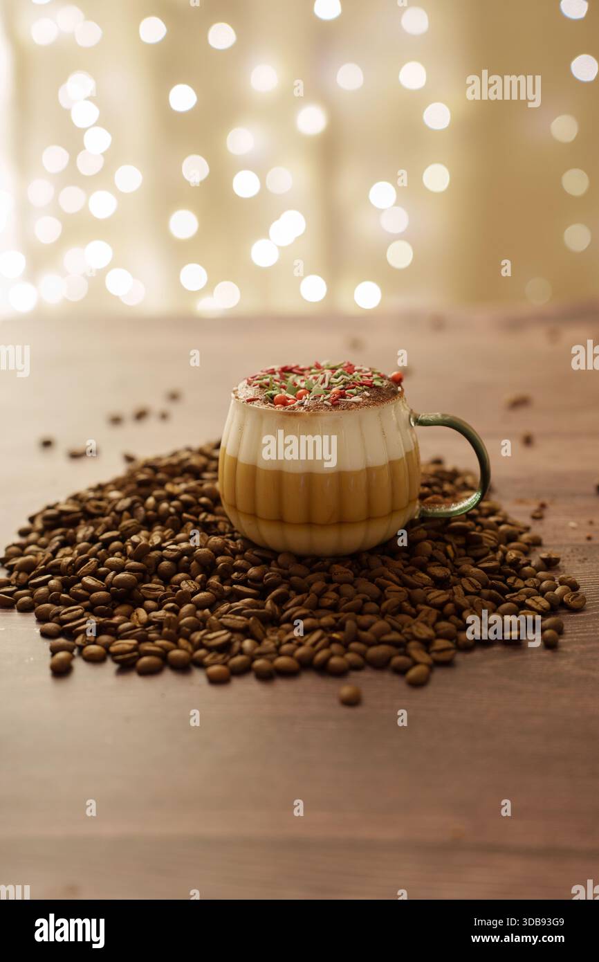 Festlicher Cappuccino in Glasbecher mit bunten Streuseln und Schaum, umgeben von Kaffeebohnen auf rustikaler Holzoberfläche mit weichem Bokeh-Licht-bac Stockfoto