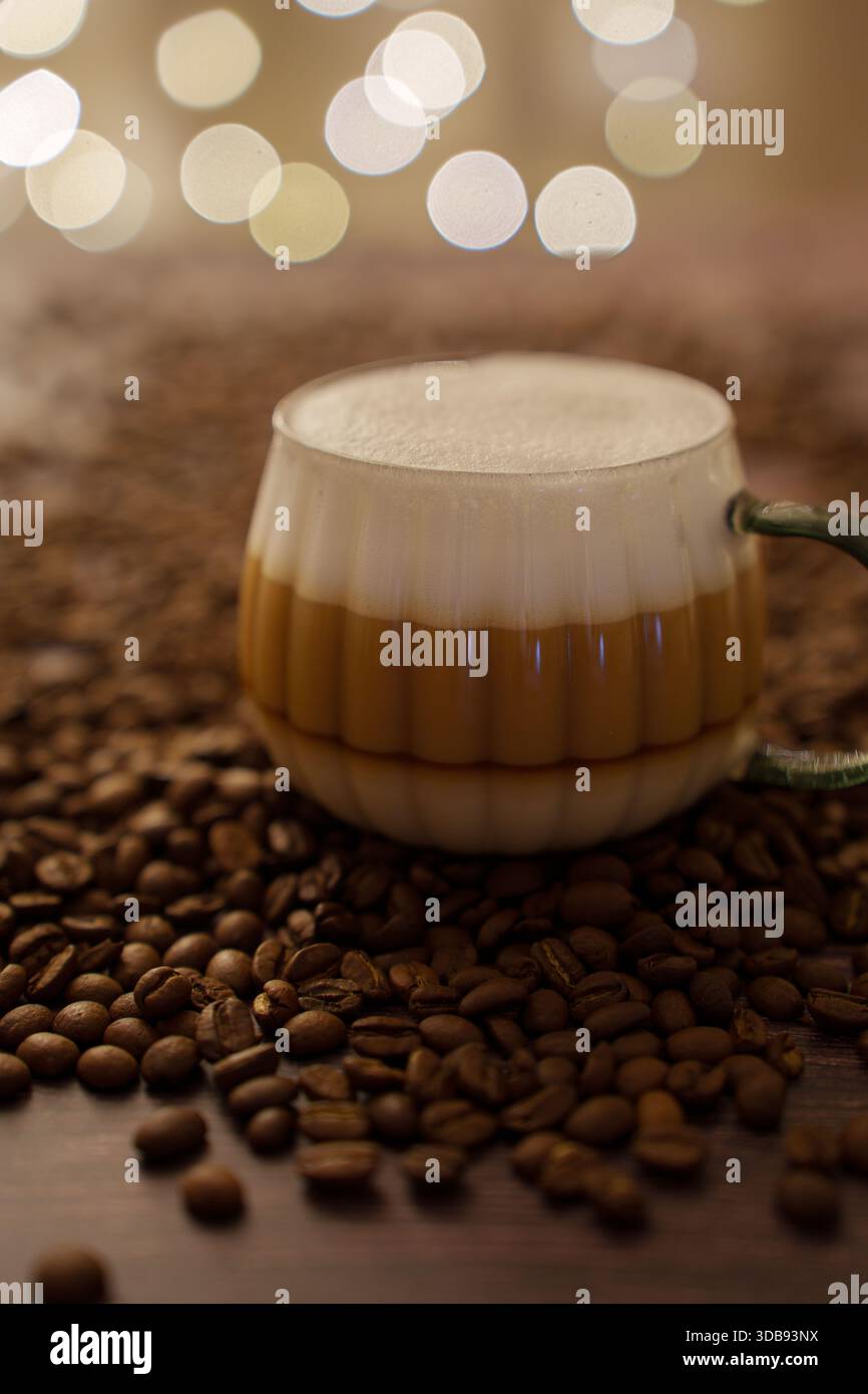 Gemütliche Szene mit geschichtetem Latte in Glasbecher, umgeben von Kaffeebohnen und warmen Bokeh-Lichtern, die eine entspannte und einladende Atmosphäre, Lifestyle und Bucht hervorrufen Stockfoto