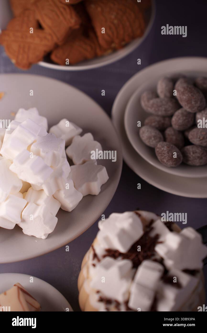 Auswahl an Snacks und Leckereien wie Marshmallows, Kekse, gezuckerte Mandeln und ein Getränk mit Schlagsahne auf weißen Tellern mit weichem Licht Stockfoto