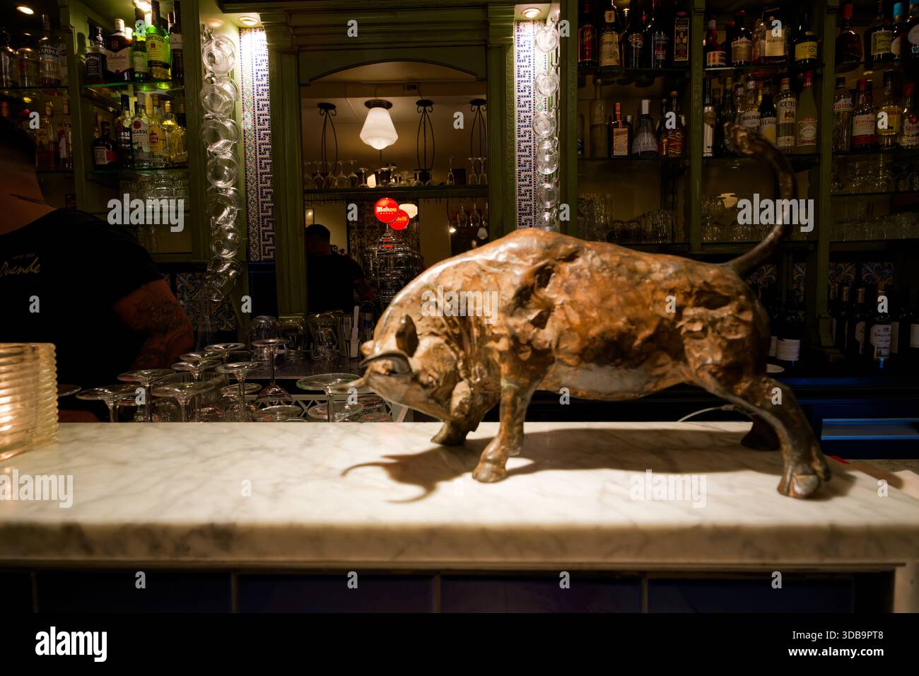 Eine Bronzestatue steht auf einer Marmortheke in einer belebten Bar mit Getränkegalons. Das warme Leuchten der Lichter verstärkt die einladende Atmosphäre Stockfoto