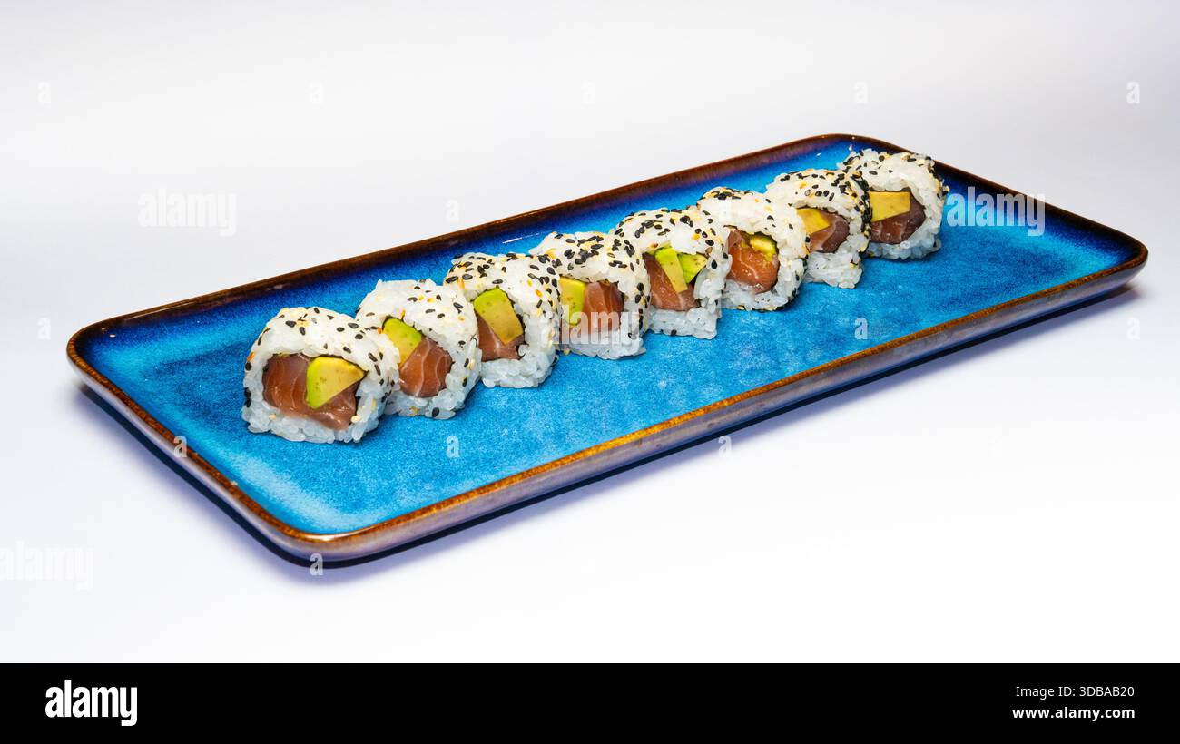Klassische Sushi-Brötchen gefüllt mit Lachs und Avocado, beschichtet mit Reis und Sesamsamen, serviert auf einer blauen Keramikplatte isoliert auf weißem Hintergrund. Stockfoto