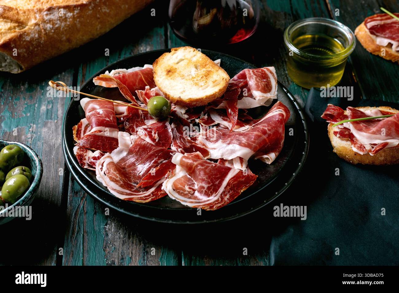 In Scheiben geschnittener spanischer Schinken Jamon Iberico paleta auf schwarzem Teller. Schüssel grüne Oliven, frisches und geröstetes Weißbrot, Olivenöl, Glas Rotwein, Tapas. Alt Stockfoto
